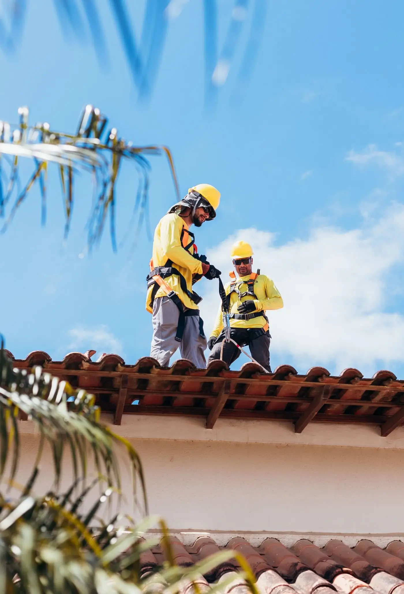 Two roofers in hard hats and safety harnesses working on a terracotta tiled roof