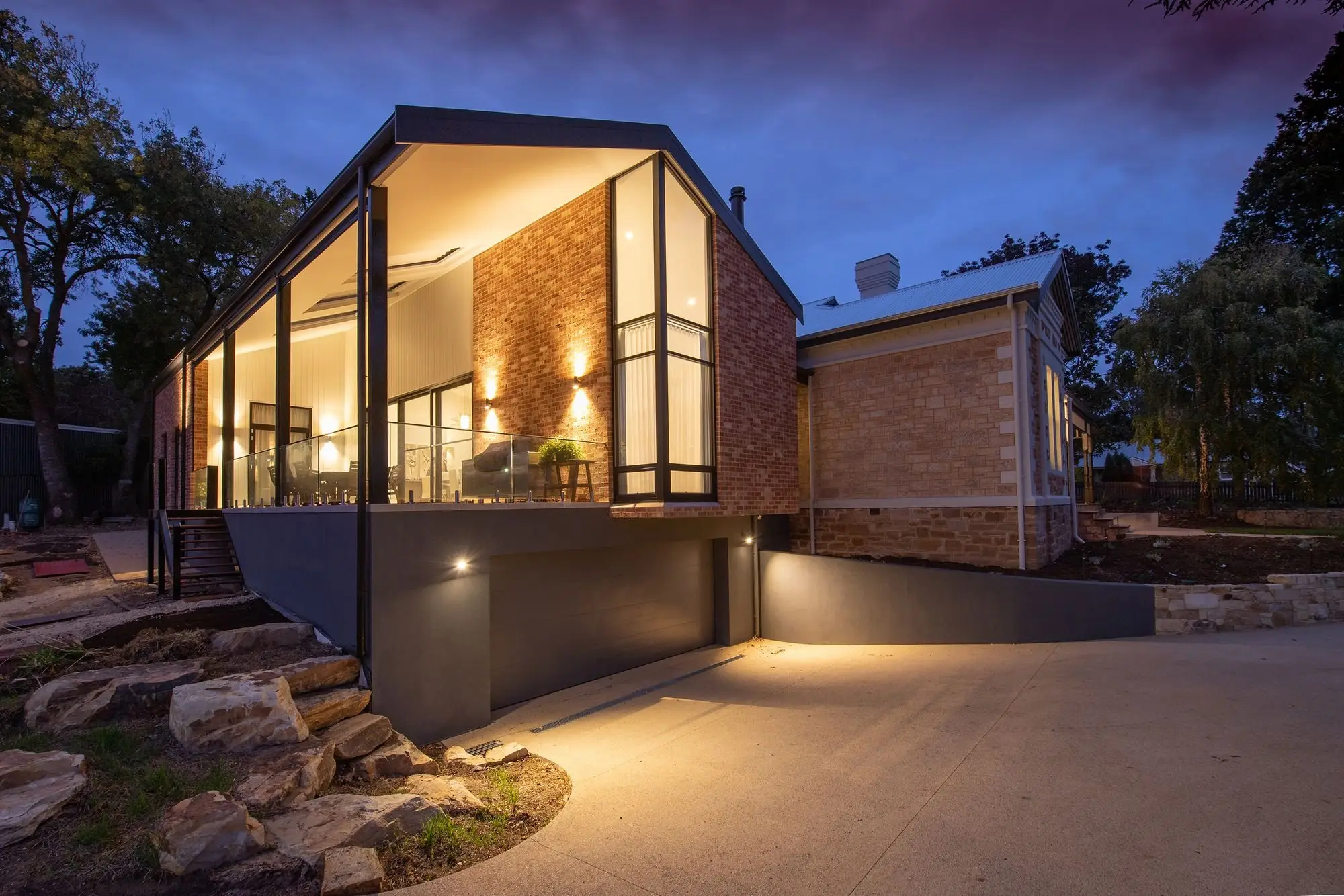 Modern gabled home at dusk with a full-height glass wall, warm-lit brick and a garage below