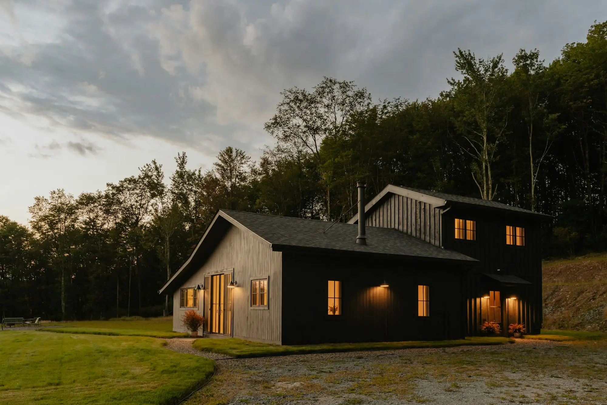 Black timber-clad house with a dark pitched roof at dusk, windows lit warm against the trees