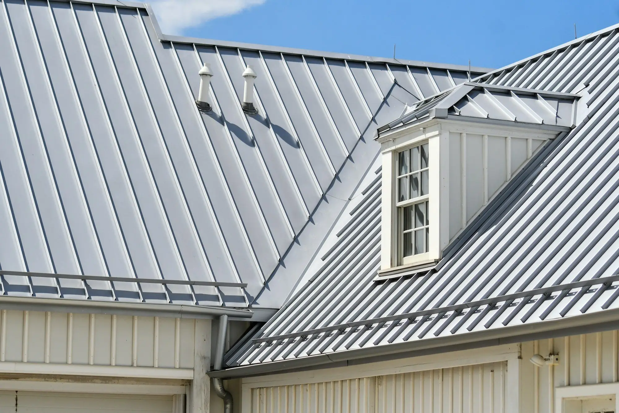 Grey standing-seam metal roof with a dormer window, crisp seams and ridge lines under a blue sky