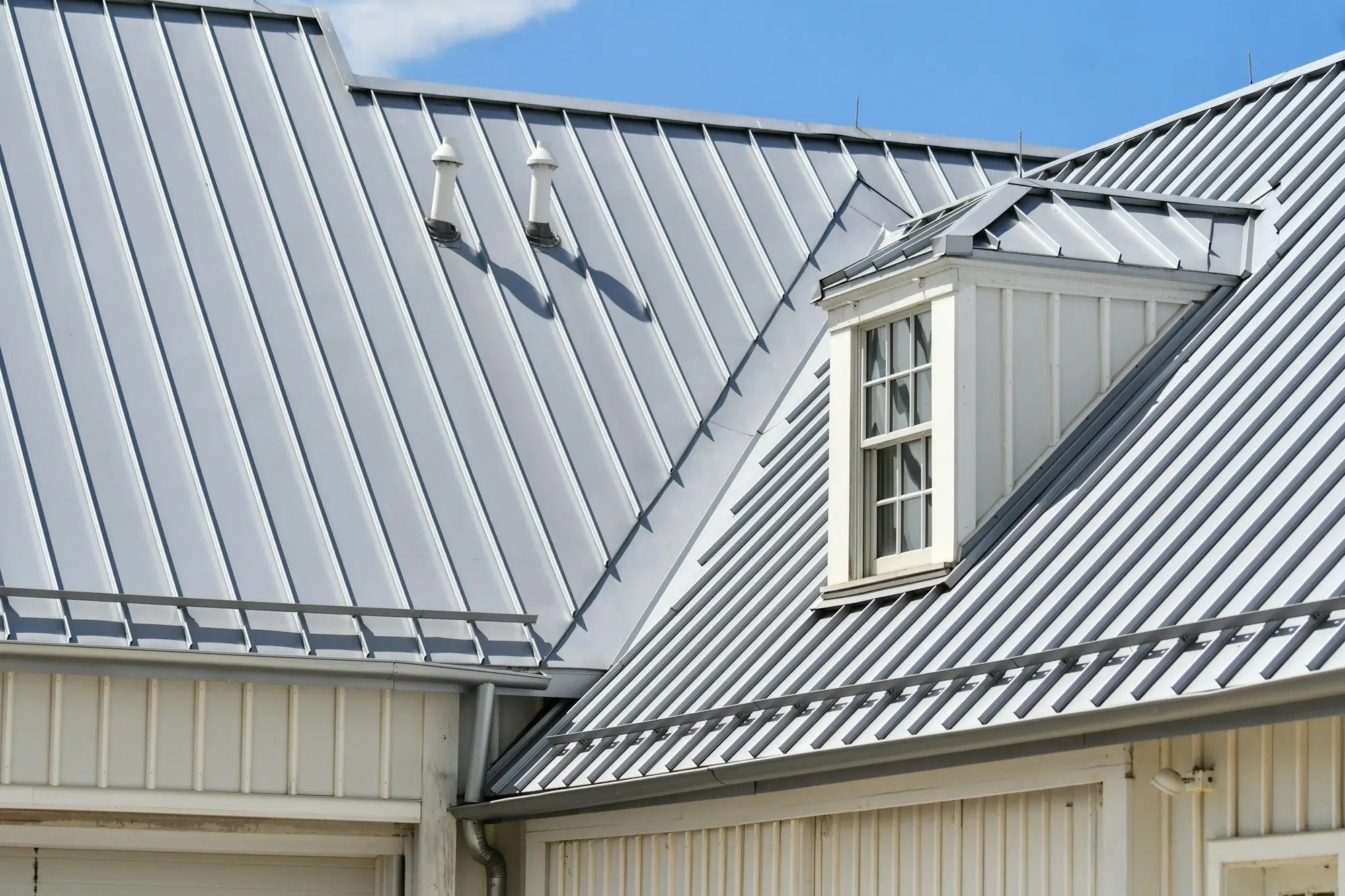 Grey standing-seam metal roof with a dormer window, crisp seams and ridge lines under a blue sky