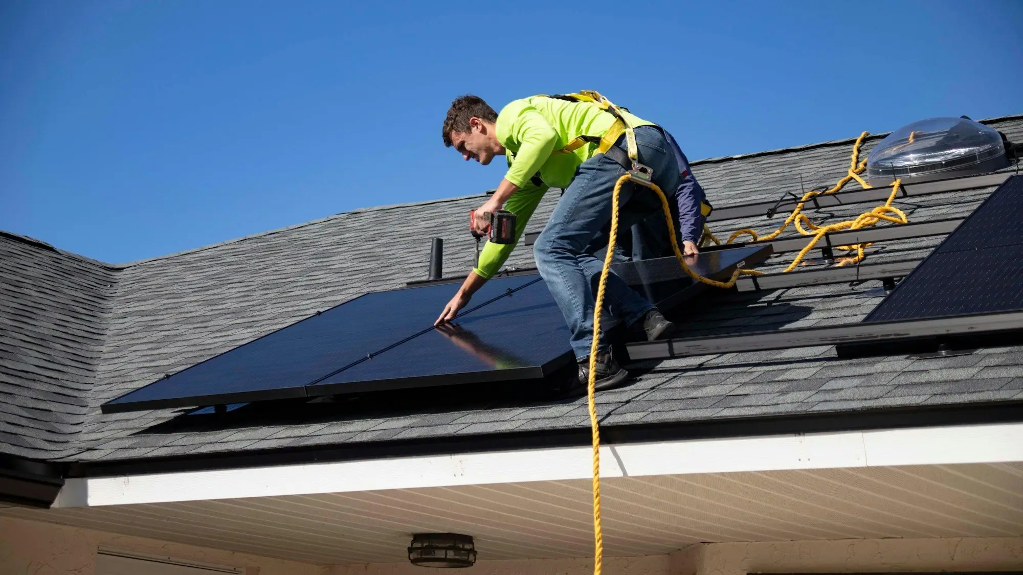 Technician in a safety harness fitting panels on a shingle roof under a clear blue sky