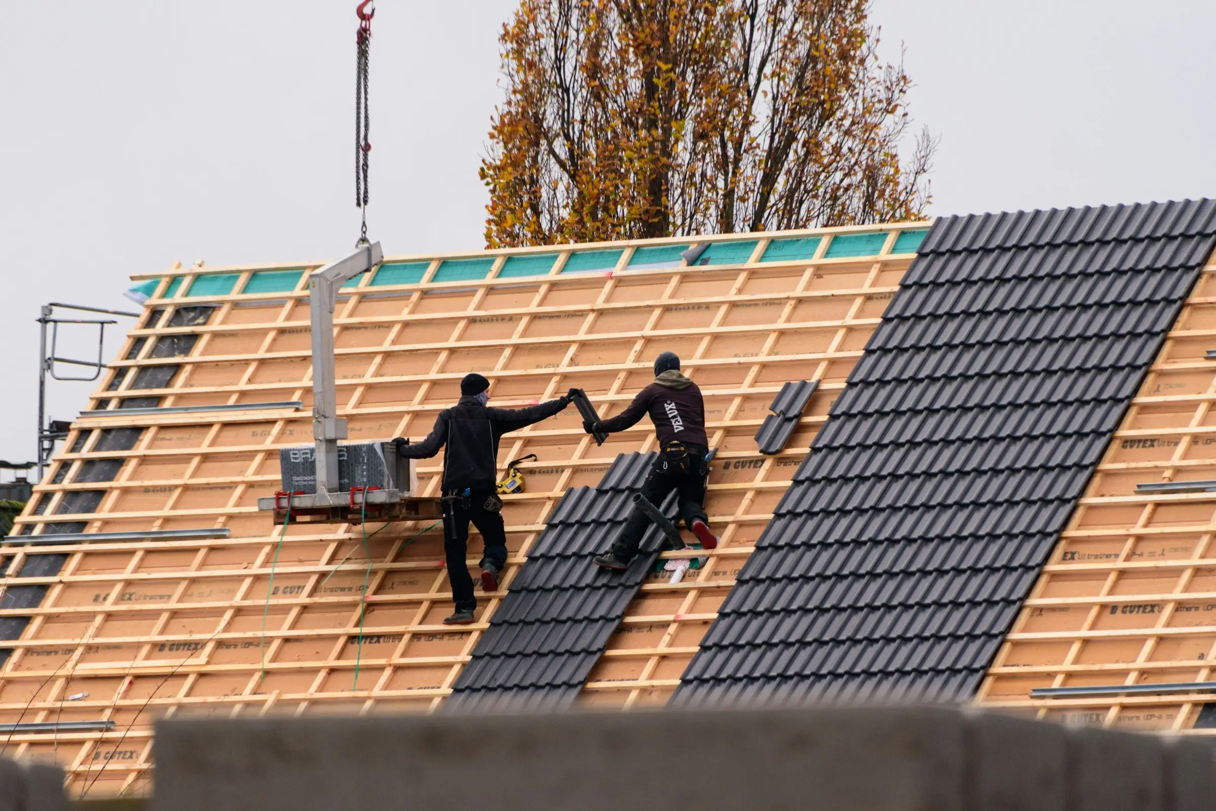 Roofers fitting dark profile roof tiles onto timber battens, with a crane lifting the next stack