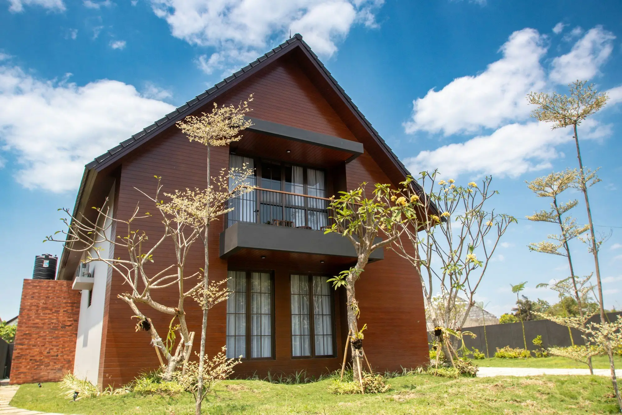 Two-storey gable-roofed home with warm timber cladding, a balcony and a garden under a blue sky