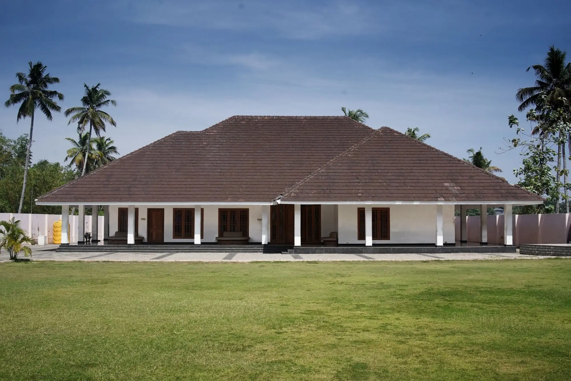 Large Kerala bungalow with a brown tiled hip roof, white pillars and a wide lawn under coconut palms