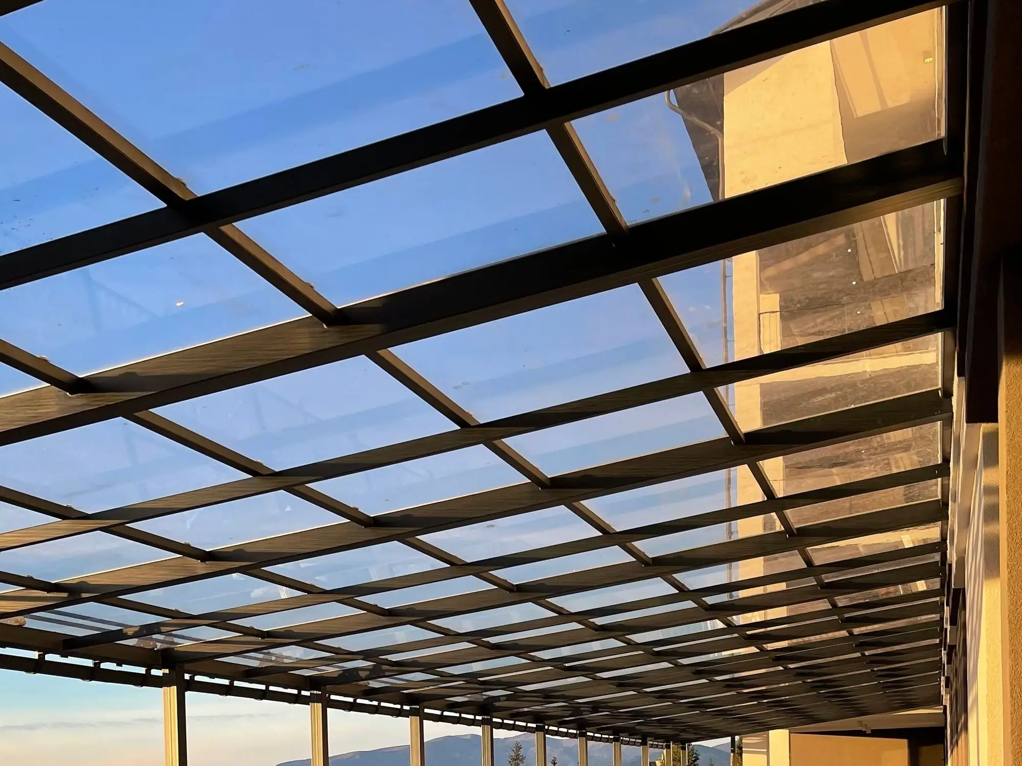 Glass canopy on a dark steel frame over a long terrace, lit by evening sun
