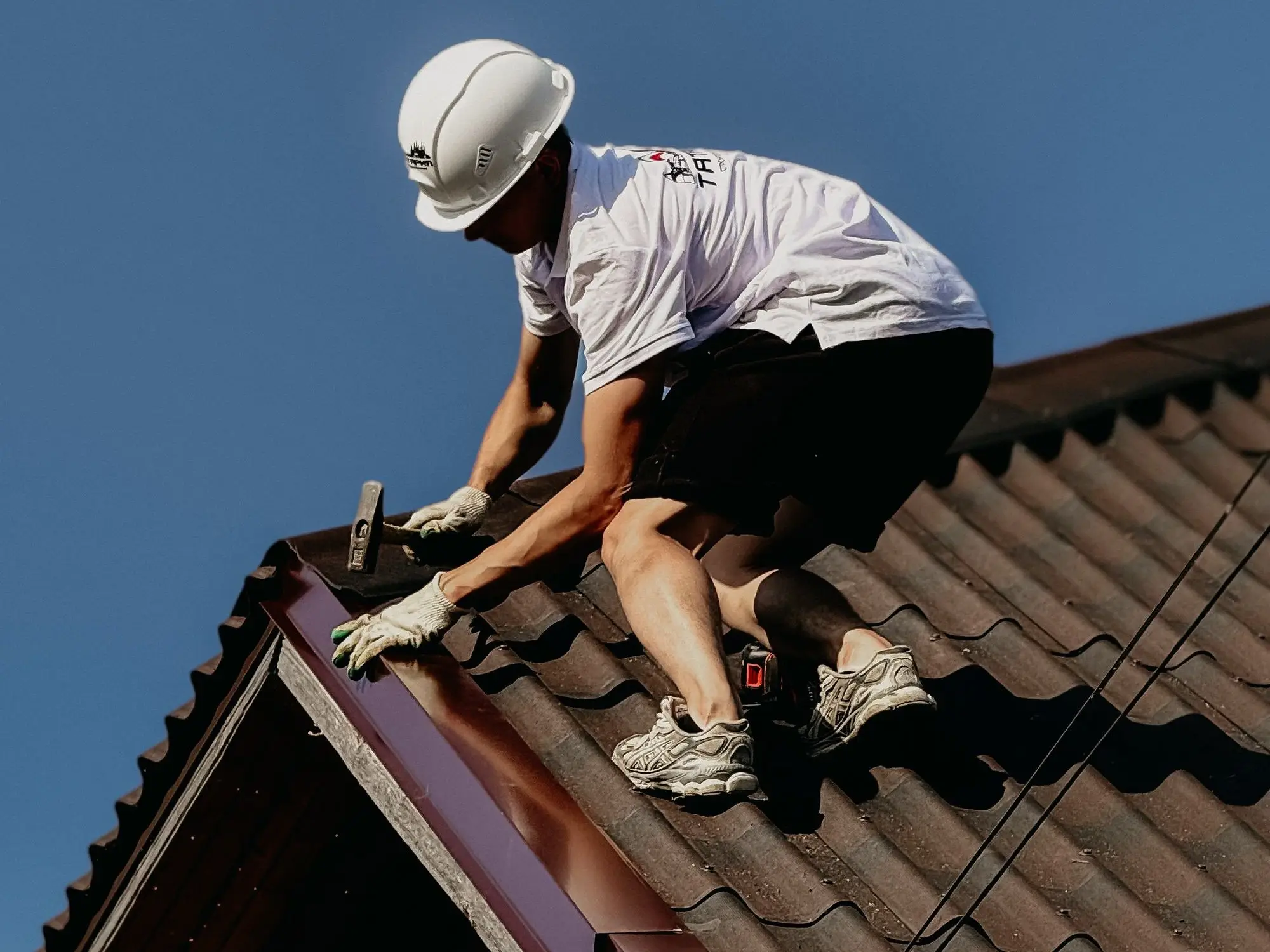 Roofer in a white hard hat and gloves fixing the ridge flashing on a dark tile-profile roof