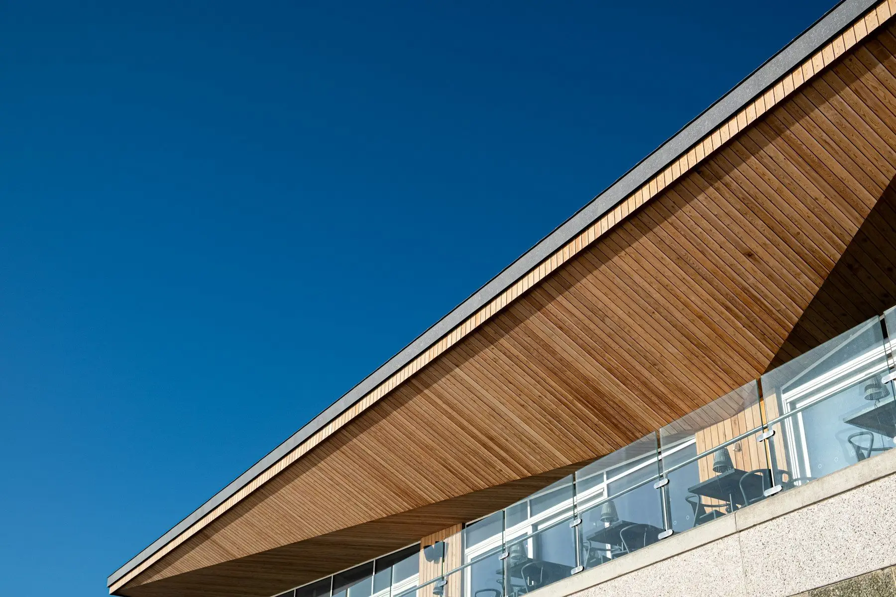 Long wood-panel soffit under a roof overhang above a glass balcony, against a blue sky